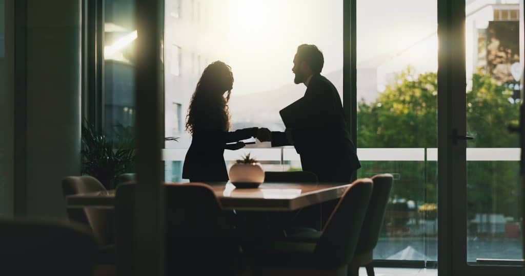 A woman is seen from a distance shaking the hand of a man in an office, with the sun shining behind them.