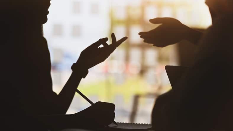 A close up of two silhouetted hands, moving in a way that suggests intimate discussion, something that can be encouraged psychological safety coaching.