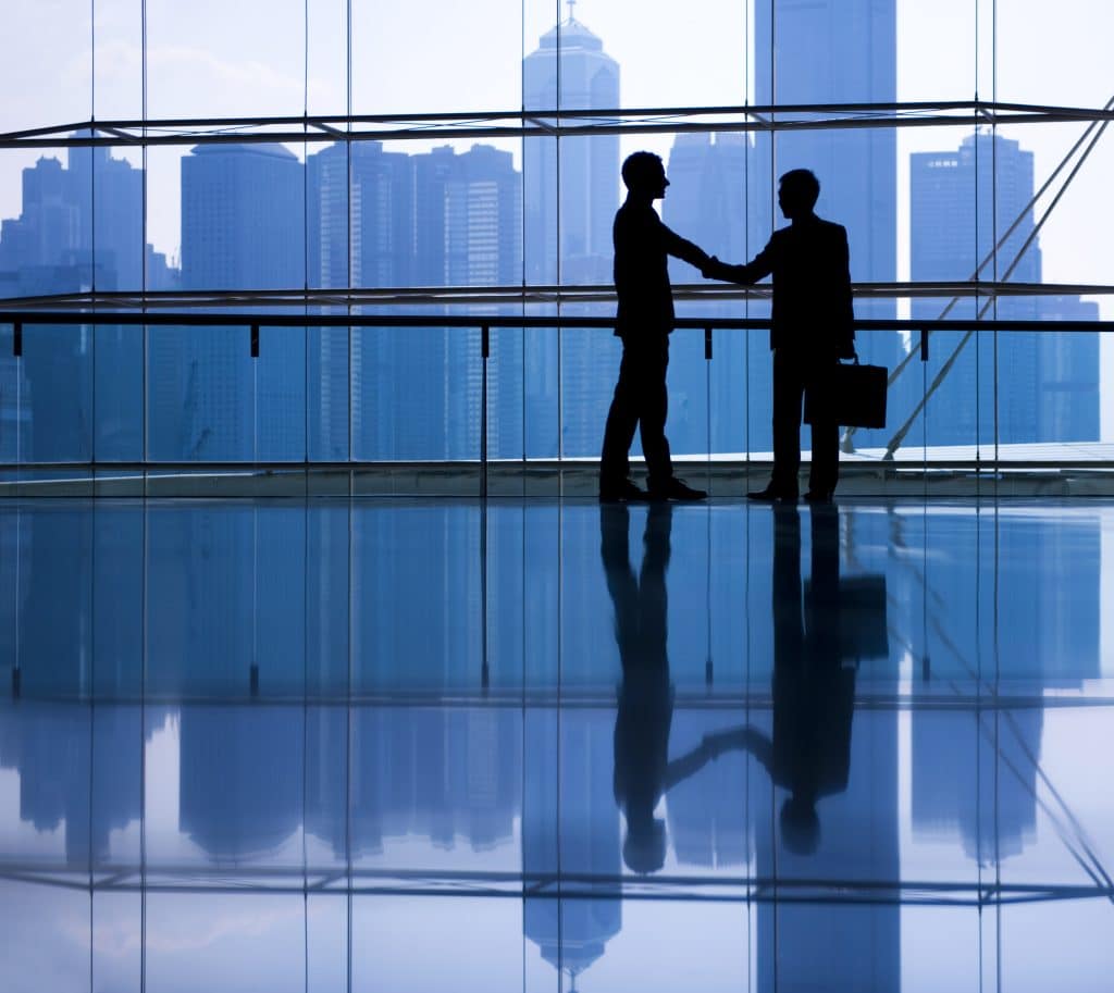 Two silhouettes of businessmen are seen standing in front of a blue, city skyline.