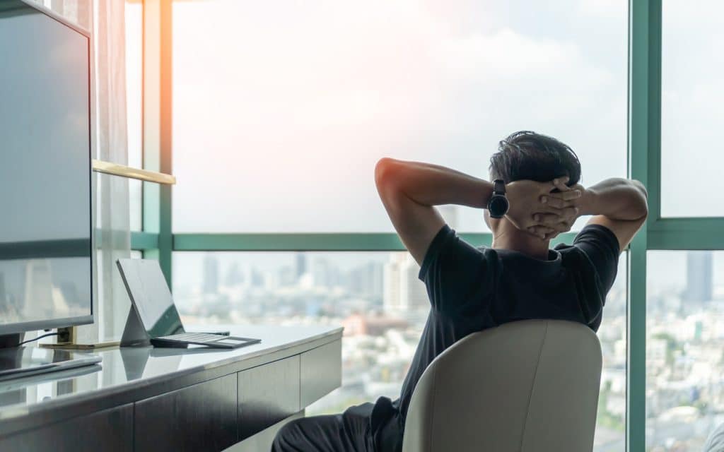 A man stares outside his window at the cityscape with his hands clasped behind his head. He's leaning back in a relaxed way, showing he has mastered work-life balance.