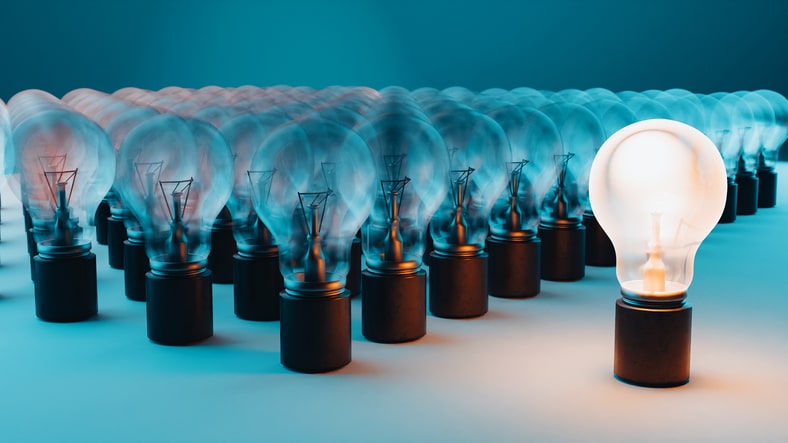 Group of light bulbs in rows, all of which are unlit except for one bulb which is glowing. The background is a gradient of blue, adding a cool tone to the scene. The glowing light bulb stands out prominently against the unlit bulbs, symbolizing concepts such as uniqueness, innovation, leadership identity, and standout ideas.