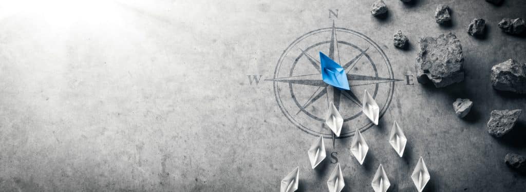 A symbolic image of paper boats, with a large blue boat leading smaller, white boats away from sharp-looking gray rocks.