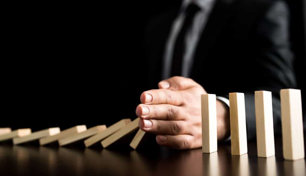 A close-up of a man in a suit, placing his hand in front of falling dominoes to prevent them from knocking over the remaining, stacked dominoes.